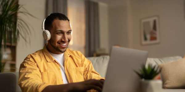 Homem sorridente com fones de ouvido em frente ao notebook durante sessão de psicoterapia online, representando brasileiros no exterior.